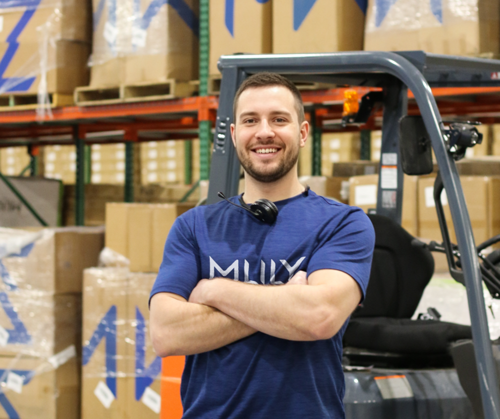 Man in a blue shirt standing in a warehouse with forklift and boxes in the background