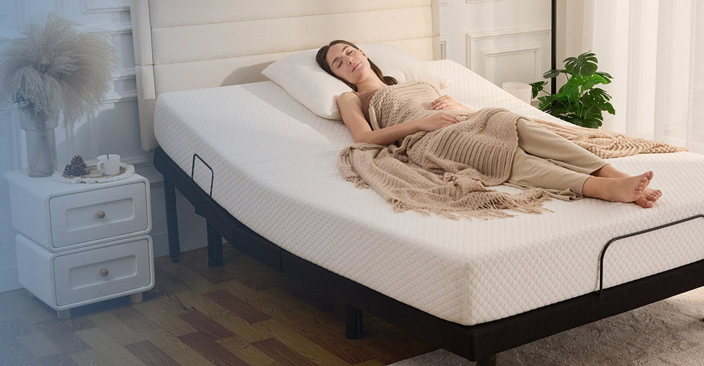 A woman lies on a white adjustable bed with a white quilted mattress, dressed in beige clothing and a fringed throw blanket in a bright bedroom setting.