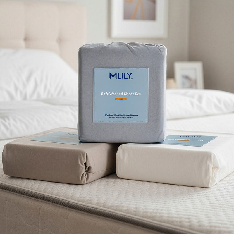 Three folded sheets in different colors (gray, beige, and off-white) with matching branded packaging labels, displayed on a bed in a softly lit bedroom setting.