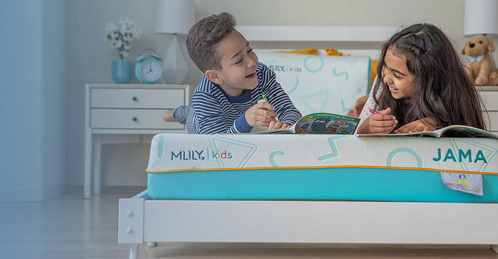 Two children reading on a mattress in a bright bedroom. The mattress is branded 'MLILY kids' with 'JAMA' written on the side, and the mattress has a white and turquoise design with a yellow accent line.