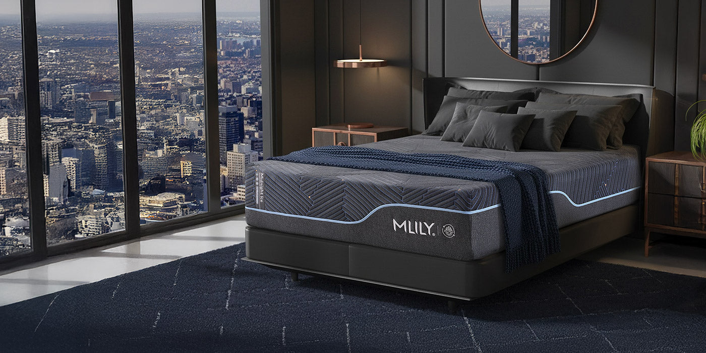 MLILY Midnight ICE mattress in a dark gray upholstered bed frame, styled in a modern bedroom with a city skyline view through large windows. The bed is dressed in gray and navy bedding with a navy throw blanket.