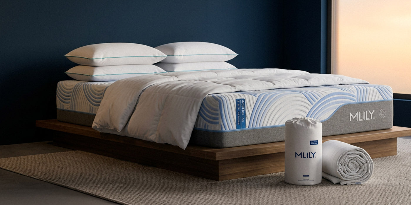 A bed dressed in white bedding with blue wave-patterned mattress and duvet, shown in a styled bedroom scene. A cylindrical and rolled white mattress protector or towel stand beside the bed, with the brand name MULLY visible on both items.