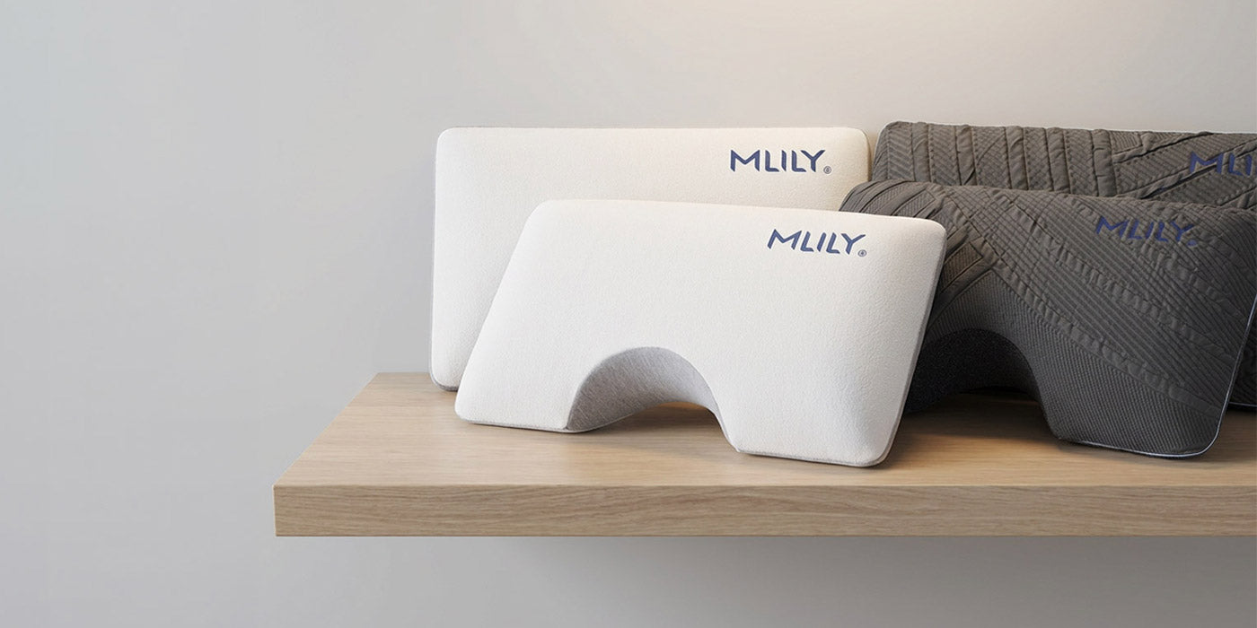 Three MLILY brand ergonomic support pillows displayed on a light wood floating shelf against a neutral light gray wall. The foreground pillow is white, with two darker gray pillows behind it.