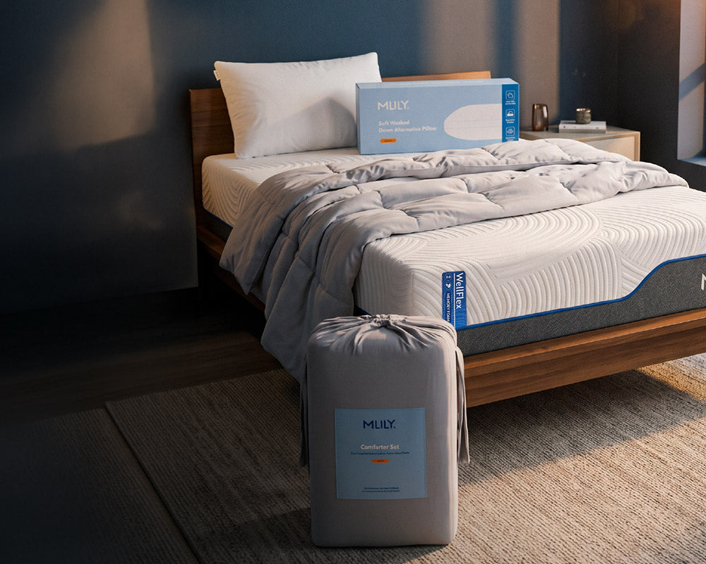 MLILY mattress shown in a styled bedroom setting, with the mattress, fitted sheet, duvet, and product box on the bed, plus a bundled mattress case in the foreground.