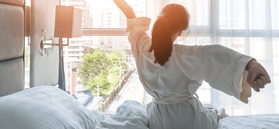 woman sits up and bed and stretches, looking toward the morning sunlight