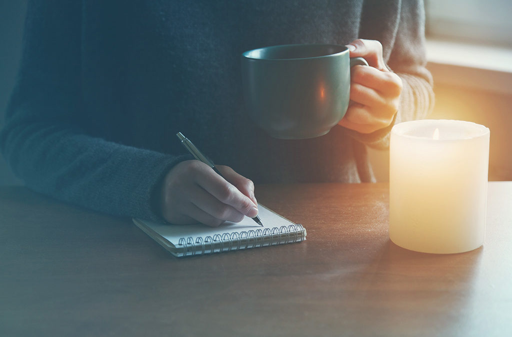 a person holds a mug and hurriedly jots some ideas on a notebook next to a candle at a table