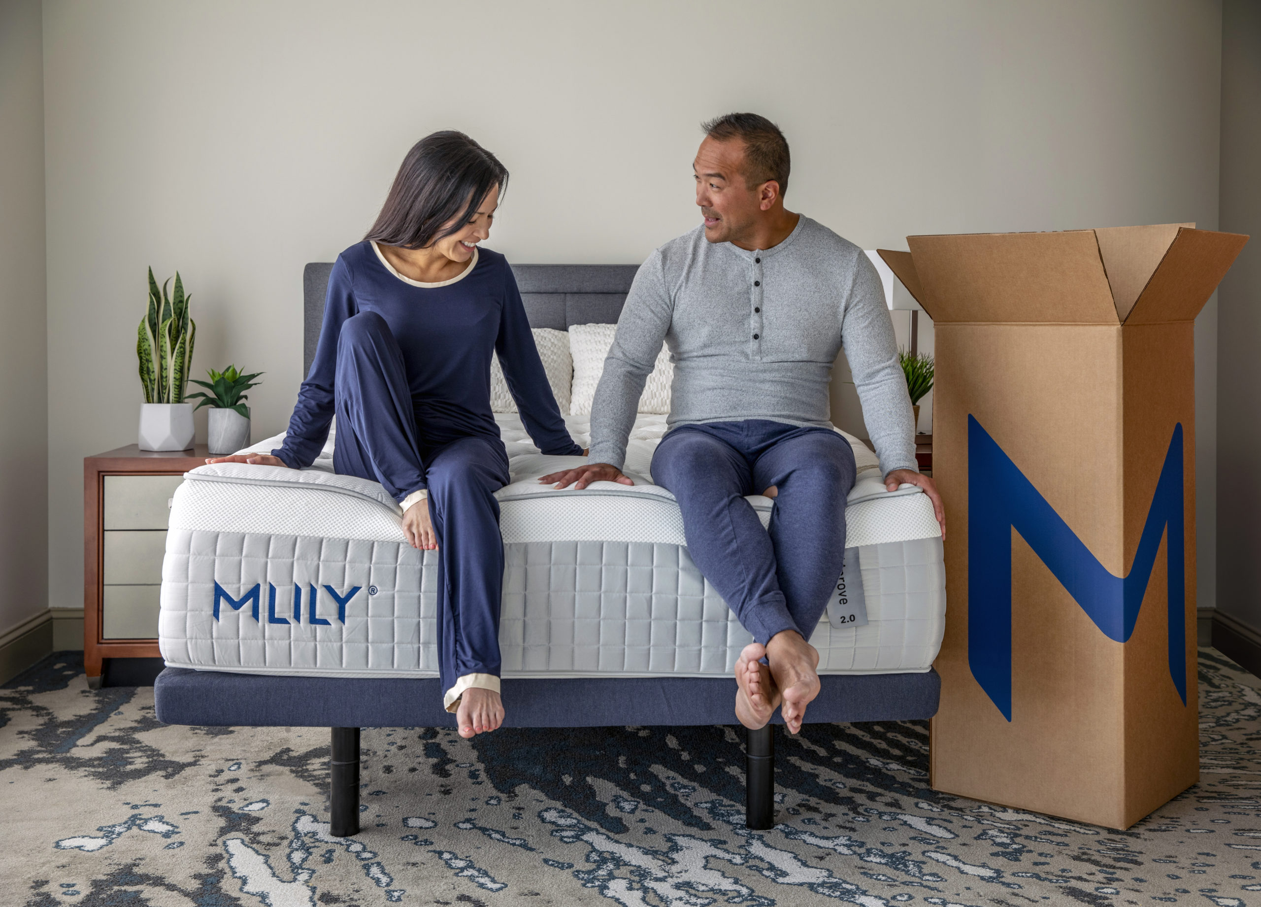 Man and woman sitting on mattress labeled MLILY on bed frame with large cardboard box next to bed.