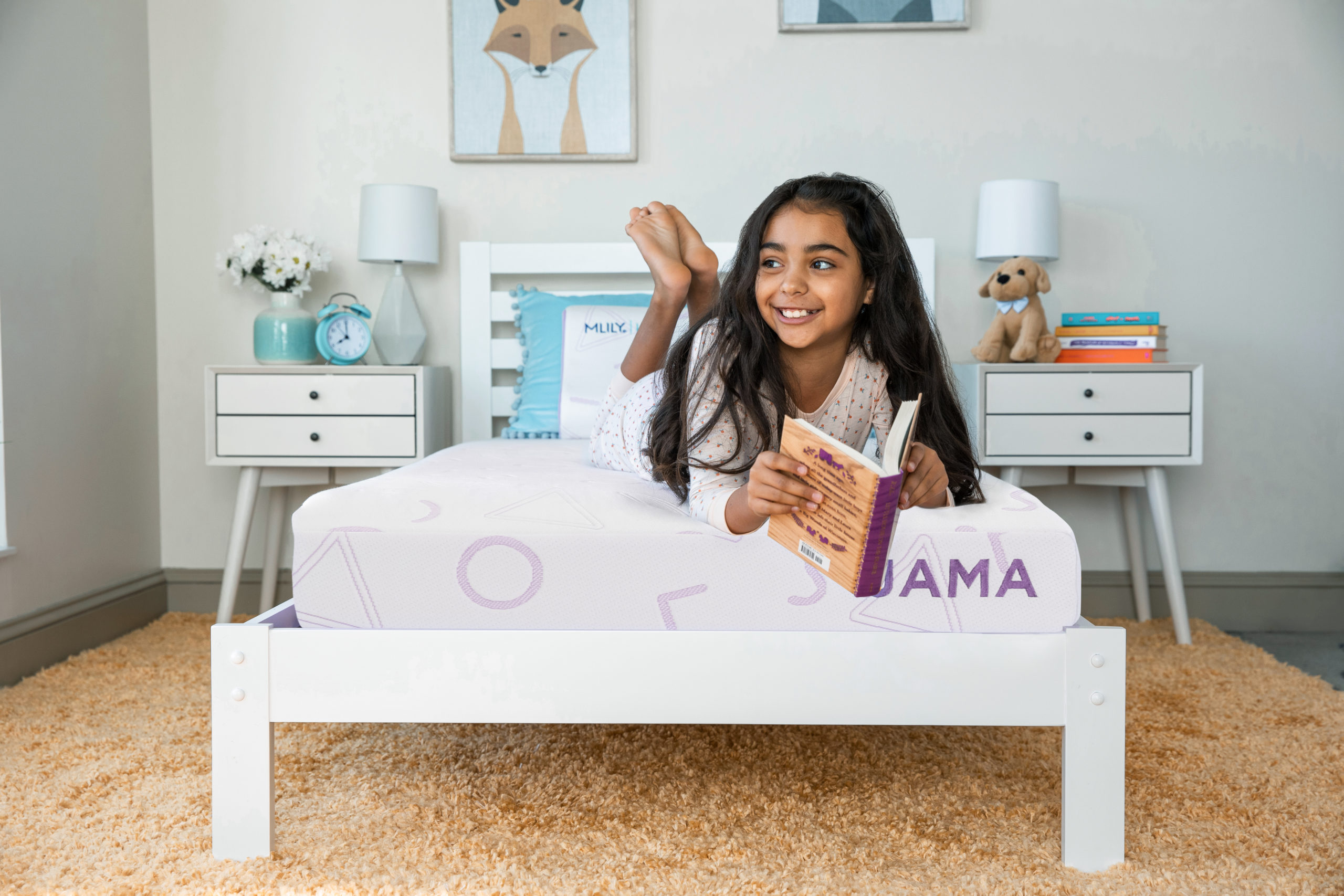 A child's bedroom with a girl laying on her stomach on a white mattress with purple designs labeled JAMA in purple, holding a book and smiling.