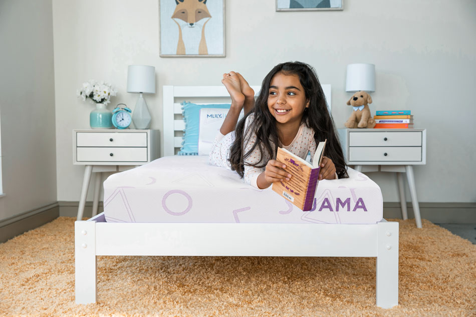 A childs bedroom featuring a girl lying on her stomach on a white mattress with purple designs labeled JAMA in purple, holding a book and smiling.