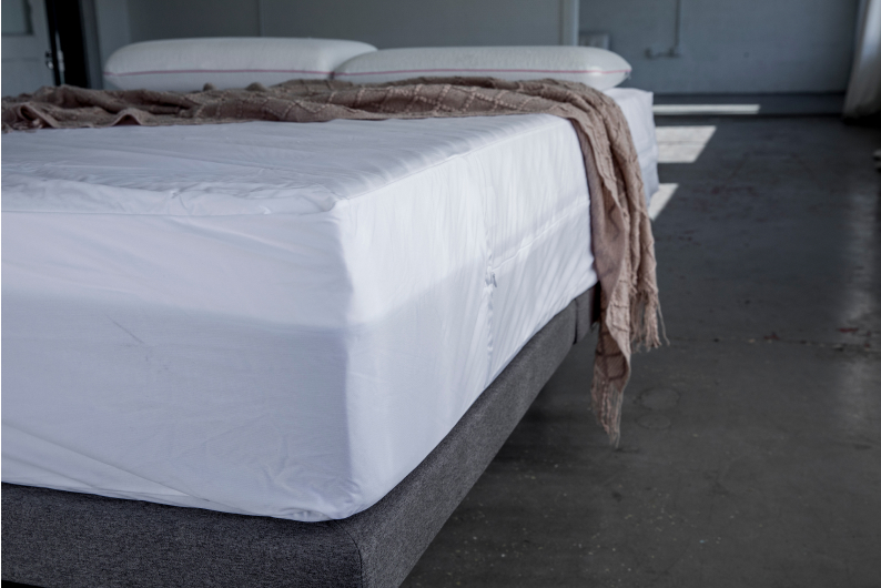 Mattress with white protector cover sitting on grey base with two pillows and a throw draped on bed.