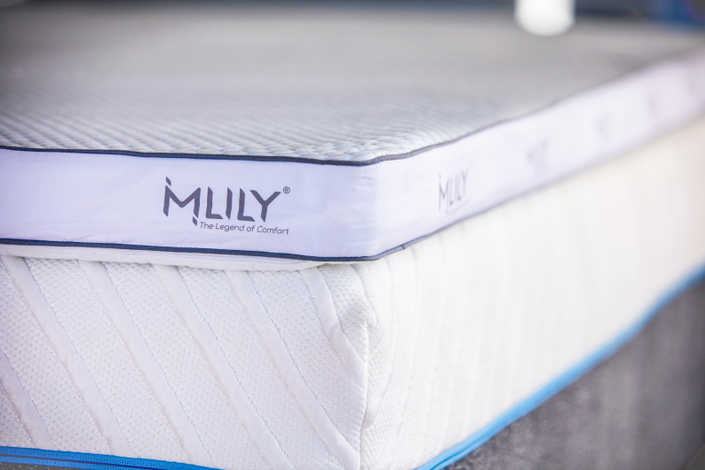 White mattress topper with navy piping labeled MLILY sitting on top of white mattress with teal piping at bottom.