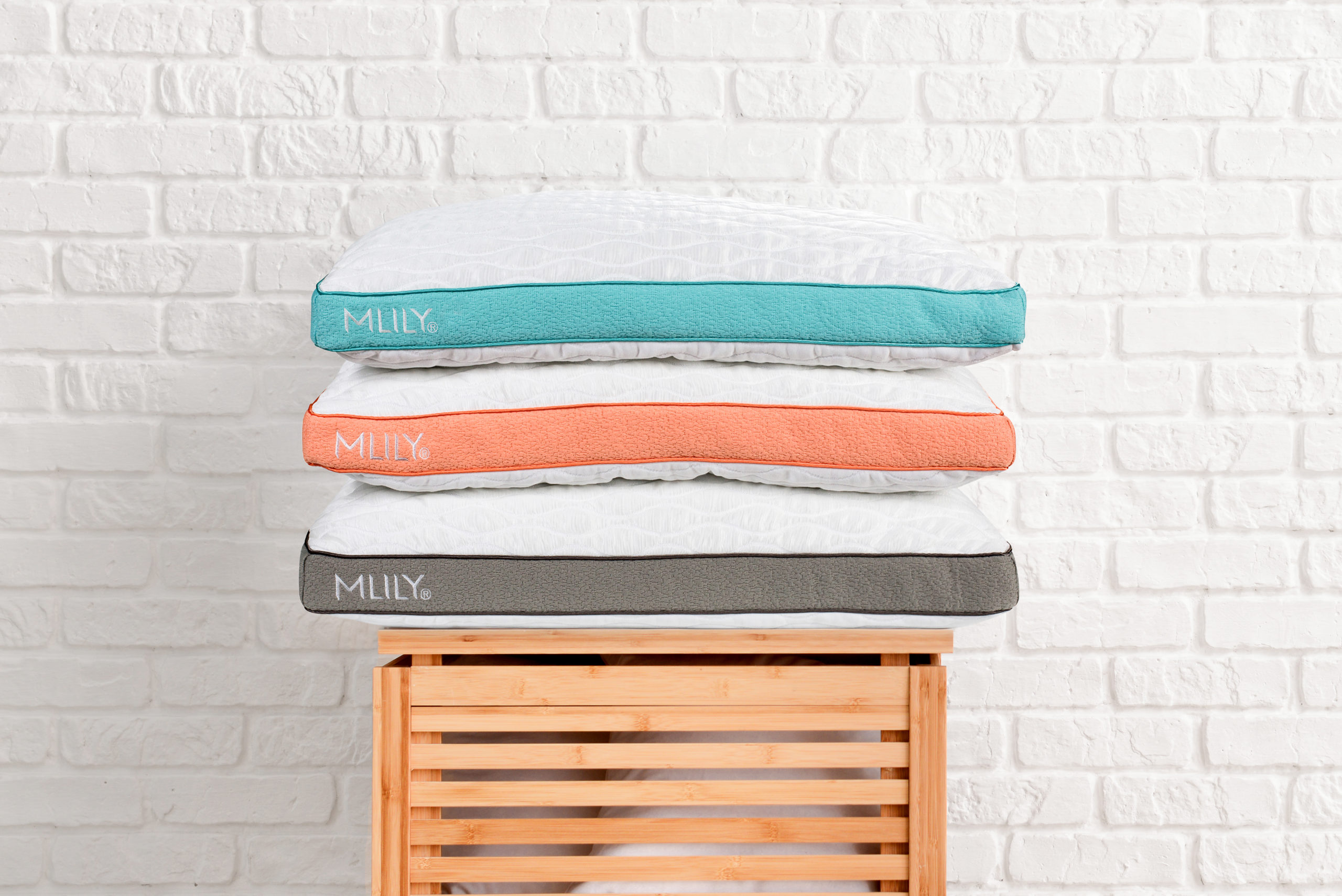 Three white MLILY pillows banded in aqua, coral, and grey stacked on a wooden slatted stool in front of white brick wall.