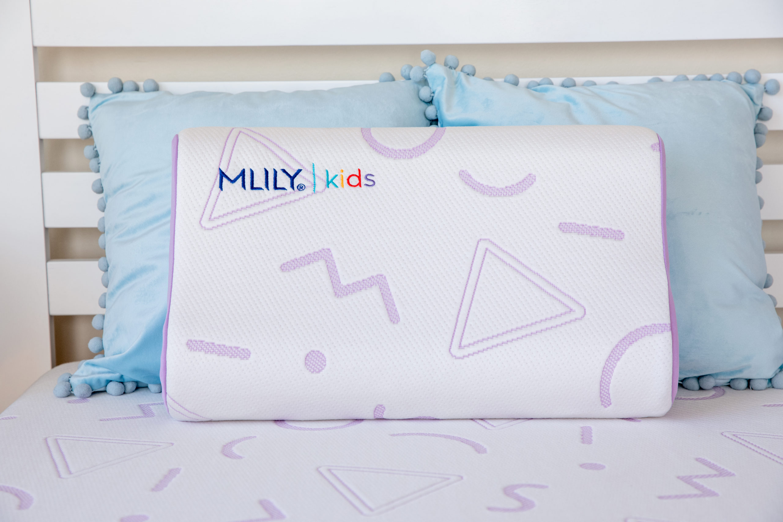 A white Jama pillow with purple accent designs labeled MLILY kids sits on a twin bed in front of two light blue throw pillows with pompom accents.