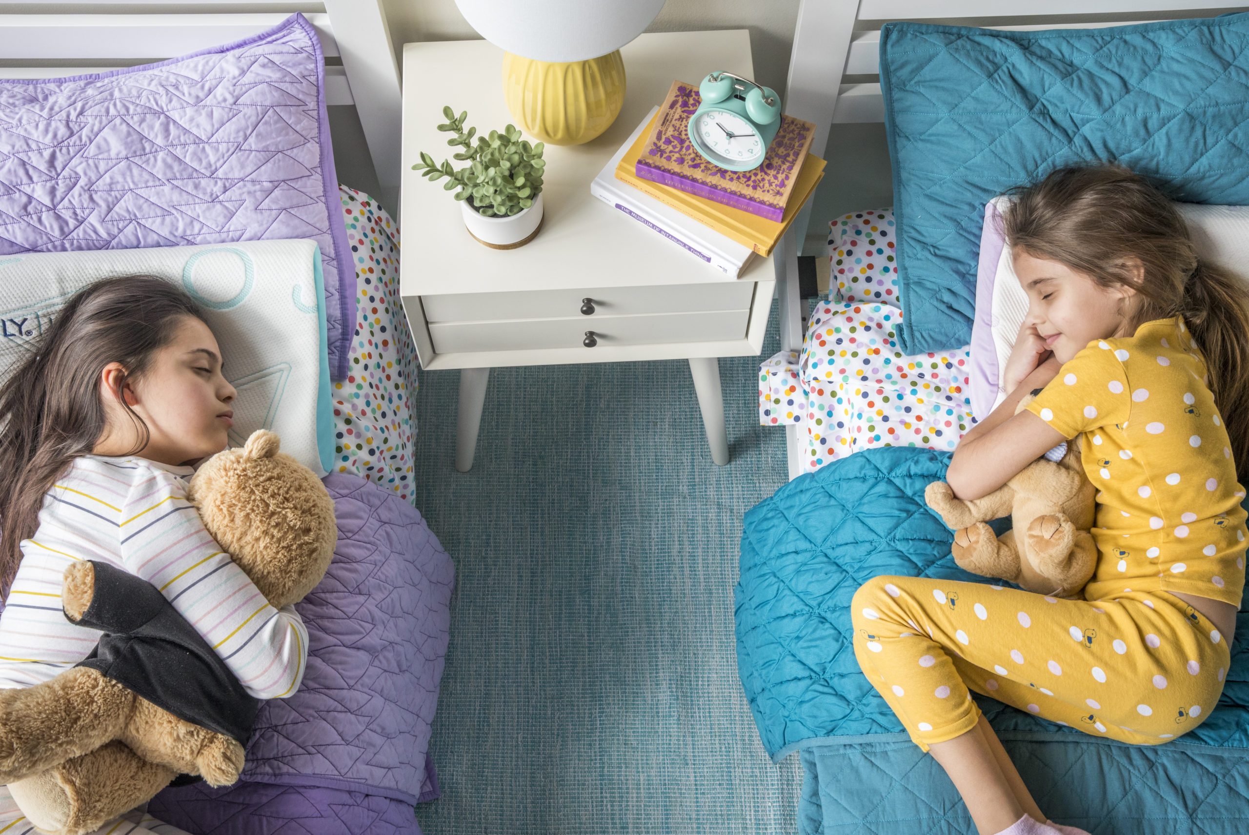 Aerial view of two girls lying in twin beds facing each other, one has purple bedding and one has teal bedding, both girls are hugging stuffed toys.