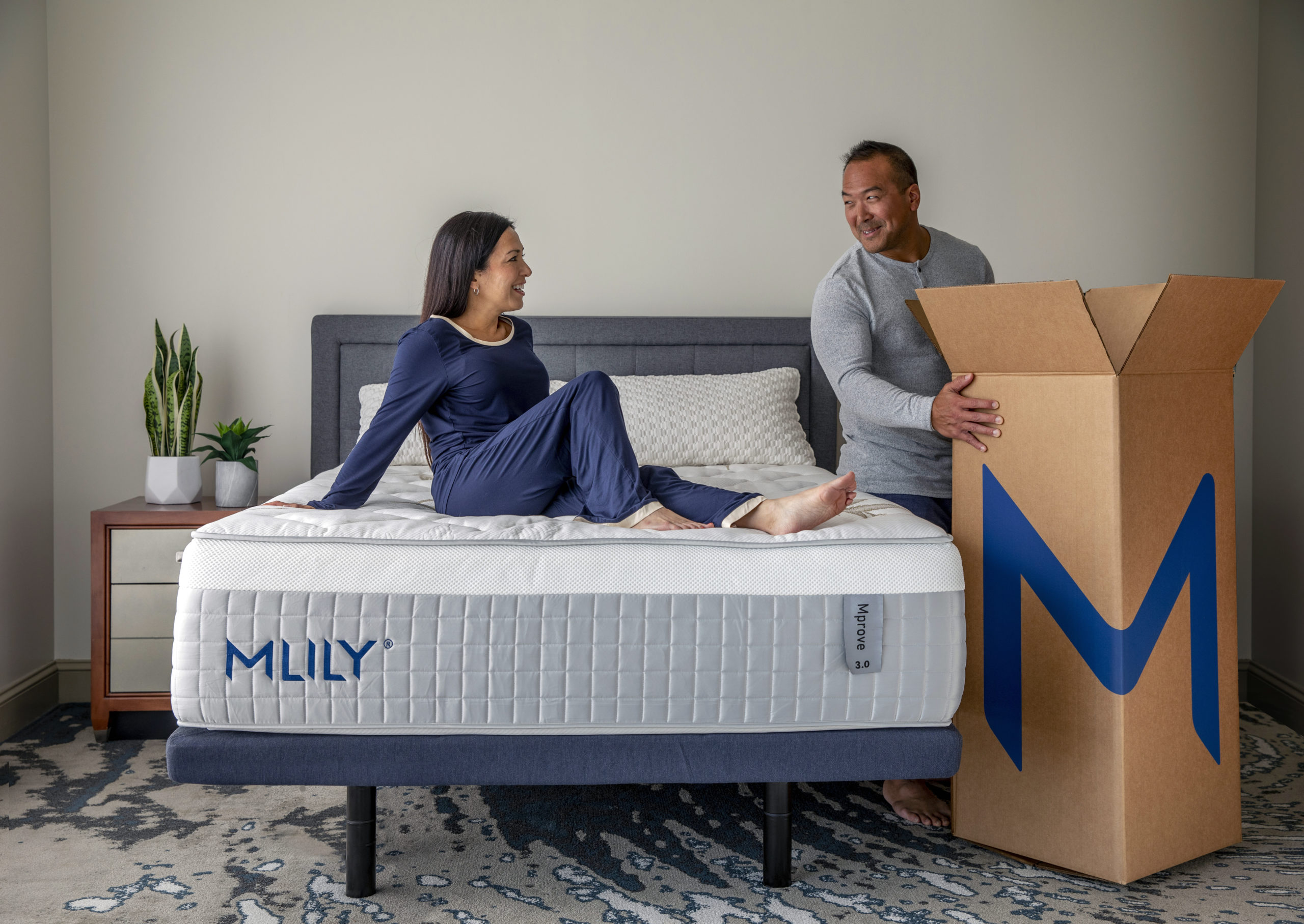 Woman in navy pajamas with dark hair reclining on MLILY-labeled mattress while looking at man in grey shirt as he holds onto large box.