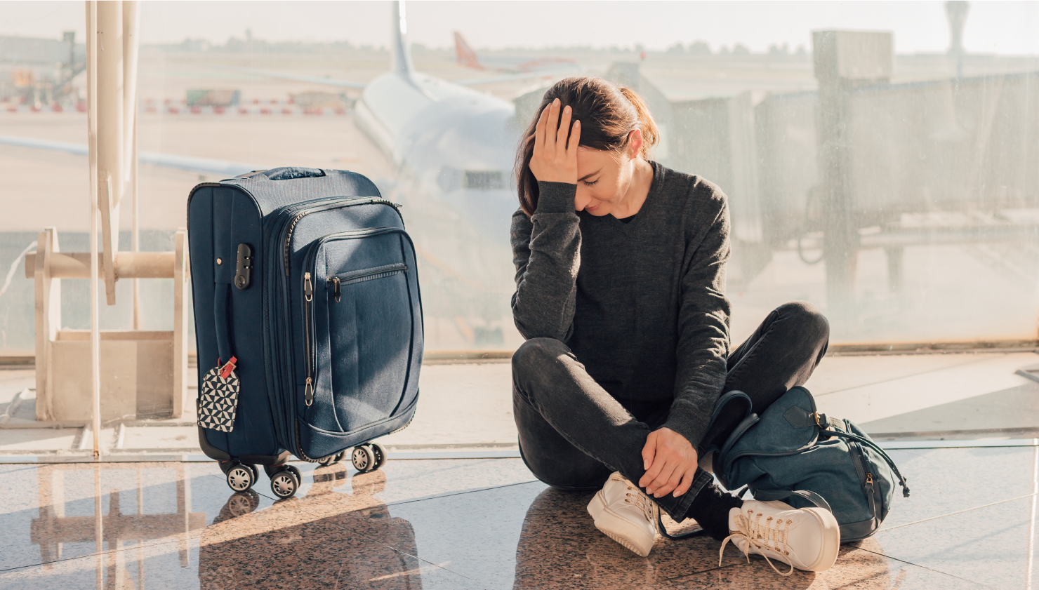 Woman in airport sitting next to suitcase seemingly in pain from jet lag