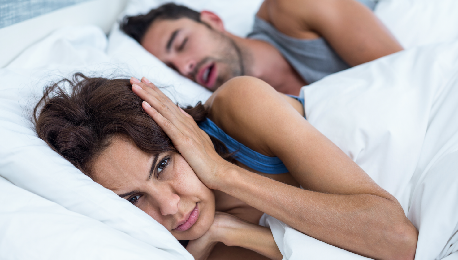 woman laying in bed covering her ears next to man snoring