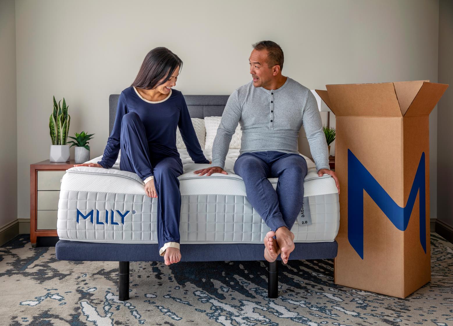Man and woman sitting on mattress labeled MLILY on bed frame with large cardboard box next to bed.
